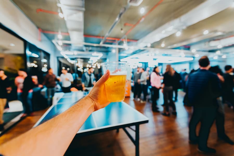 A cold lager held up over a ping pong table, the room crowded behind it